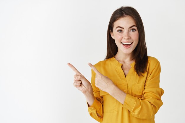 Podekscytowana młoda kobieta pokazująca baner, wskazująca palcem w lewo i uśmiechająca się do kamery, stojąca zdumiona na białej ścianie