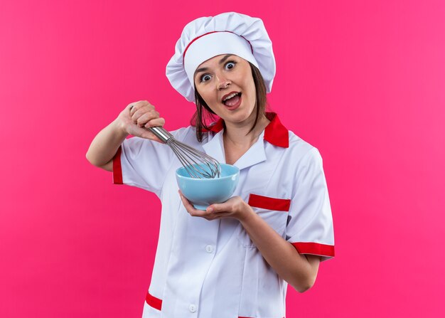 Podekscytowana młoda kucharka ubrana w mundur szefa kuchni trzymająca miskę z trzepaczką odizolowaną na różowej ścianie