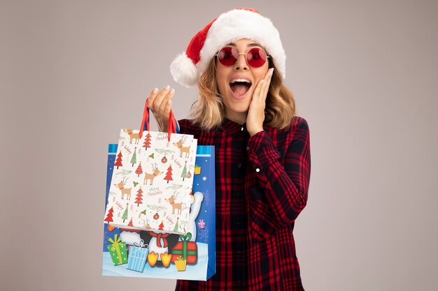 Podekscytowana młoda piękna dziewczyna w świątecznym kapeluszu w okularach, trzymająca torby z prezentami, kładąc rękę na policzku na białym tle