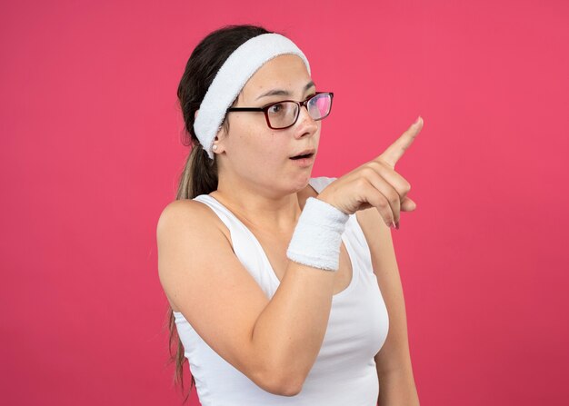 Podekscytowana młoda sportowa dziewczyna w okularach optycznych nosząca opaskę i opaski na nadgarstki wygląda i wskazuje z boku