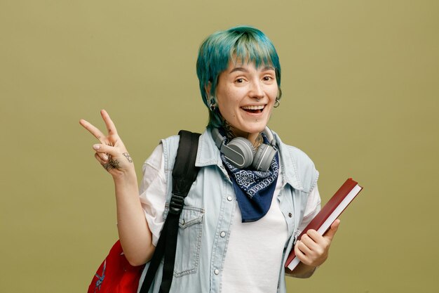 Podekscytowana młoda studentka nosząca słuchawki i chustkę na szyi i plecaku trzymająca notatnik patrząc na kamerę pokazującą znak pokoju na białym tle na oliwkowym tle