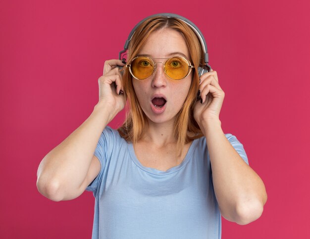 Podekscytowana ruda dziewczyna imbir z piegami w okularach przeciwsłonecznych i na słuchawkach, patrząc na kamery na różowo