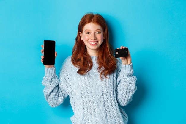 Podekscytowana ruda dziewczyna pokazuje ekran telefonu komórkowego i kartę kredytową, pokazując sklep internetowy lub aplikację, stojąc na niebieskim tle.
