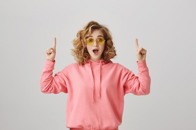 Podekscytowana Stylowa Dziewczyna Kręcone, Wskazując Palcami W Górę, Aby Pokazać Reklamę