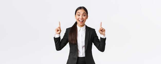 Podekscytowana uśmiechnięta azjatycka sprzedawczyni w garniturze sugeruje wiele wskazując palcem w górę, mówiąc szczegóły Kobieta ogłasza ogłoszenie i pokazuje górny baner na białym tle