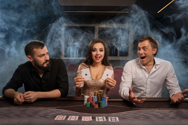 Podekscytowani przyjaciele grają w pokera w kasynie z dymem, ciesząc się z wygranej i patrząc w kamerę. Młodzi stawiają zakłady, czekając na ogromną wygraną. Hazard za pieniądze. Gry losowe.