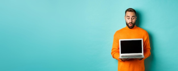 Bezpłatne zdjęcie podekscytowany dorosły model męski pokazujący ekran laptopa demonstrujący promocję online stojącą szczęśliwie przeciwko lig