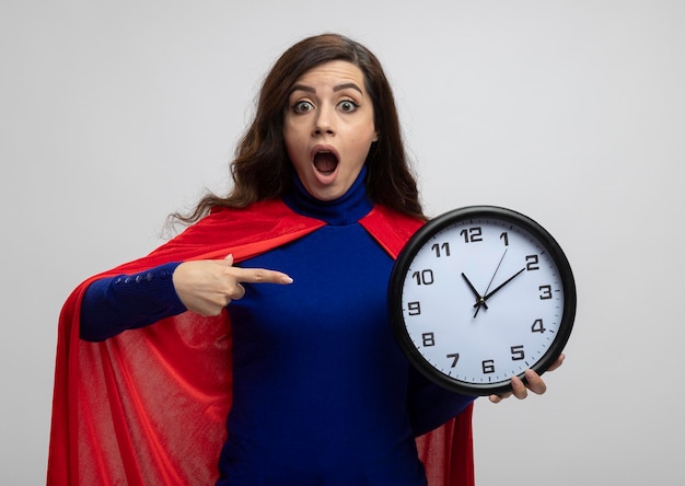 Podekscytowany kaukaski dziewczyna superbohatera z czerwoną peleryną trzyma i wskazuje na zegar na białym tle