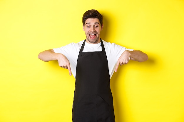 Podekscytowany kelner w czarnym fartuchu, wskazujący palcami w dół, sprawdzający ofertę promocyjną, stojący na żółtym tle.