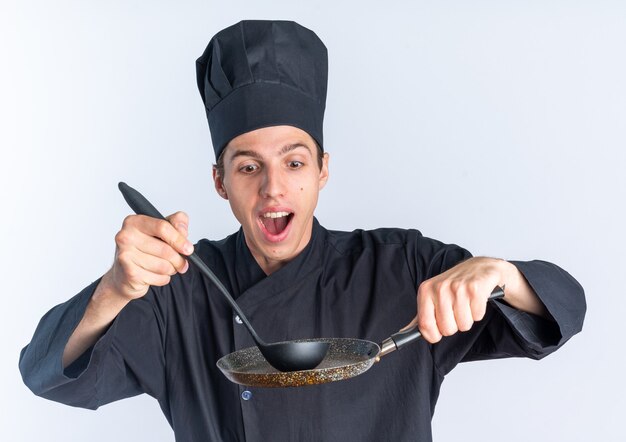 Podekscytowany młody blond mężczyzna kucharz w mundurze szefa kuchni i czapce dotykając patelni z kadzią, patrząc na to na białym tle na białej ścianie