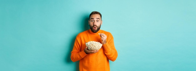 Podekscytowany Młody Człowiek Oglądający Ciekawy Film Na Ekranie Telewizora, Jedzący Popcorn I Wyglądający Na Zdumionego Blue Bac