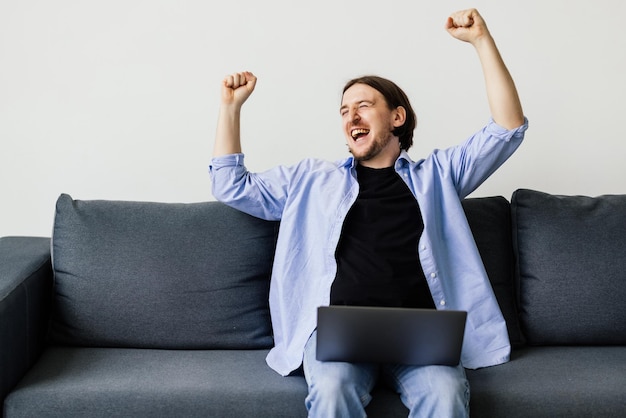 Podekscytowany młody człowiek wygrywa używając laptopa siedząc na kanapie w domu