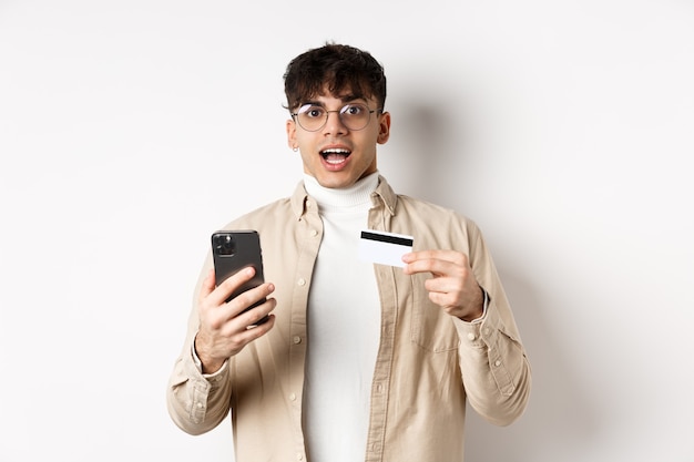 Podekscytowany młody człowiek zakupy online, trzymając telefon komórkowy i plastikową kartę kredytową, dokonując zakupu w internecie, stojąc na białym tle.