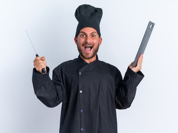 Podekscytowany młody kaukaski kucharz w mundurze szefa kuchni i czapce trzymającej tasak i deskę do krojenia, patrząc na kamerę na białym tle na białej ścianie