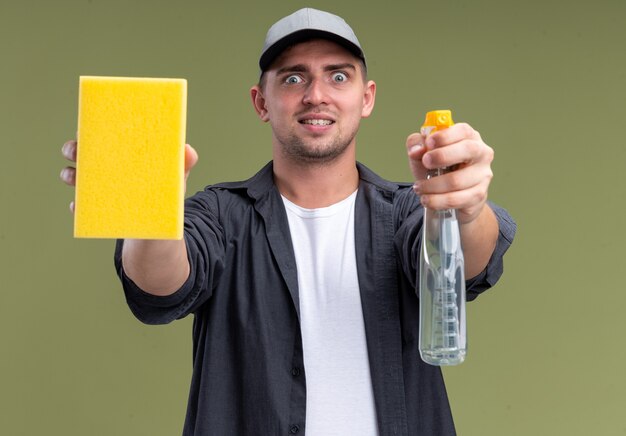 Podekscytowany młody przystojny facet sprzątający na sobie t-shirt i czapkę, wyciągający gąbkę z butelką z rozpylaczem na białym tle na oliwkowej ścianie