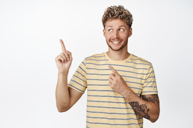 Podekscytowany przystojny młody mężczyzna uśmiechający się wskazując i patrząc szczęśliwy na lewy górny róg pustej przestrzeni pokazującej logo marki sprzedaży stojącej na białym tle