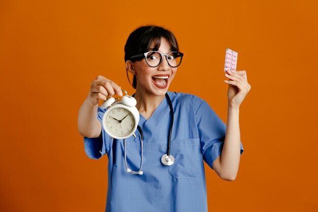 Podekscytowany trzymając budzik z pigułkami młoda lekarka nosi jednolity stetoskop fith na białym tle na pomarańczowym tle