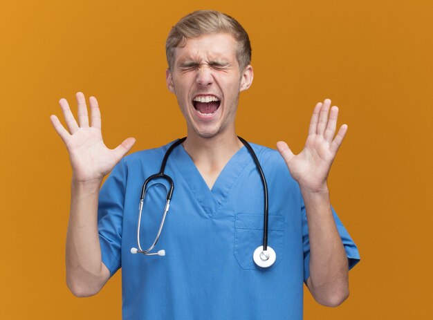 Podekscytowany z zamkniętymi oczami młody mężczyzna lekarz ubrany w mundur lekarza ze stetoskopem, rozkładając ręce na białym tle na pomarańczowej ścianie