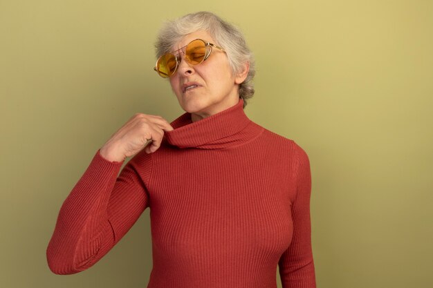 Podgrzana stara kobieta w czerwonym swetrze z golfem i okularach przeciwsłonecznych, ciągnąca za kołnierz swetra z zamkniętymi oczami odizolowanymi na oliwkowozielonej ścianie z kopią przestrzeni