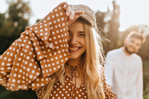 Pogodna dziewczyna śmieje się patrząc na kamerę na tle swojego chłopaka Kobieta w czapce i topie w groszki pozuje w parku