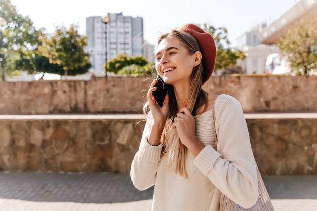 Pogodna dziewczyna z długimi włosami rozmawia przez telefon na ulicy. Atrakcyjna biała kobieta w berecie podczas zabawy na świeżym powietrzu.