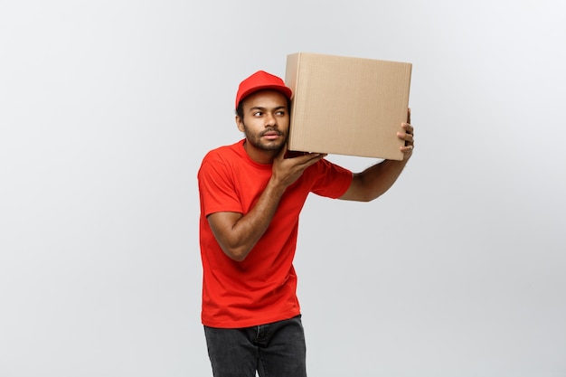 Pojęcie dostawy - Portret Ciekawych Afro-amerykański mężczyzna dostawy słuchać wewnątrz opakowania pudełko. Pojedynczo na tle Szarym Studio. Skopiuj miejsce.