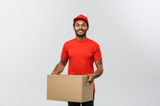 Pojęcie dostawy - Portret Szczęśliwego Afroamerykanów dostawy mężczyzna w czerwonym szmatką gospodarstwa pakiet pudełko. Pojedynczo na tle Szarym Studio. Skopiuj miejsce.