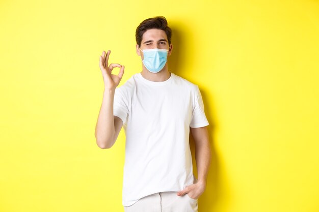 Pojęcie koronawirusa, pandemii i dystansu społecznego. Pewny siebie młody człowiek w masce medycznej pokazuje dobrze znak i mrugając, żółte tło.