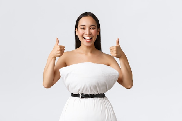 Pojęcie piękna, mody i mediów społecznościowych. Szczęśliwa uśmiechnięta azjatycka kobieta bawi się biorąc udział w internetowym wyzwaniu poduszkowym, robiąc sukienkę z poduszki i pasa owijającego się wokół odpadów, pokaż kciuki do góry