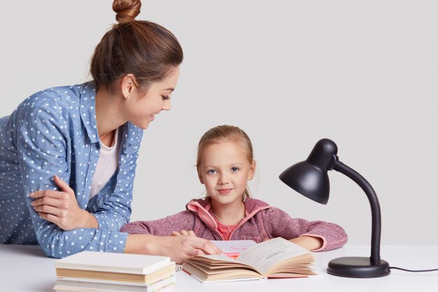 Bezpłatne zdjęcie pojęcie rodzicielstwa, nauki i edukacji, niebieskookie dziecko siedzi w miejscu pracy, czyta książkę wraz z matką, uczy się wiersza na pamięć, pozuje w przytulnym pokoju na białym