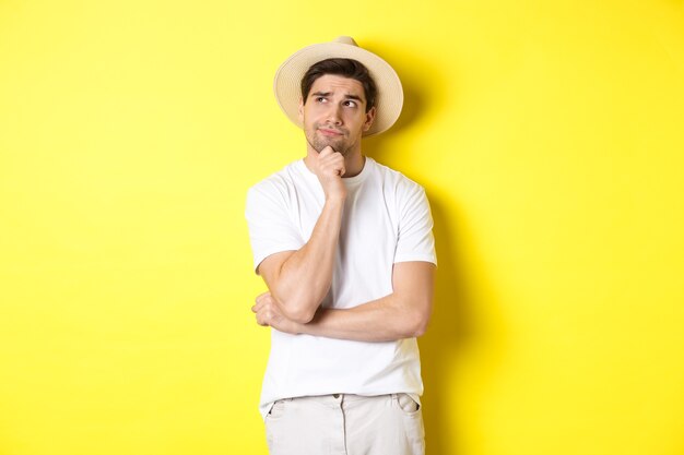 Pojęcie turystyki i lata. Zamyślony turysta mężczyzna, patrząc w lewy górny róg i myślący, stojący w słomkowym kapeluszu i białej koszulce na żółtym tle.