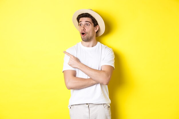 Pojęcie turystyki i stylu życia. Podekscytowany przystojny facet w słomkowym kapeluszu sprawdzający reklamę, wskazujący i patrząc na logo w lewym górnym rogu, żółte tło.
