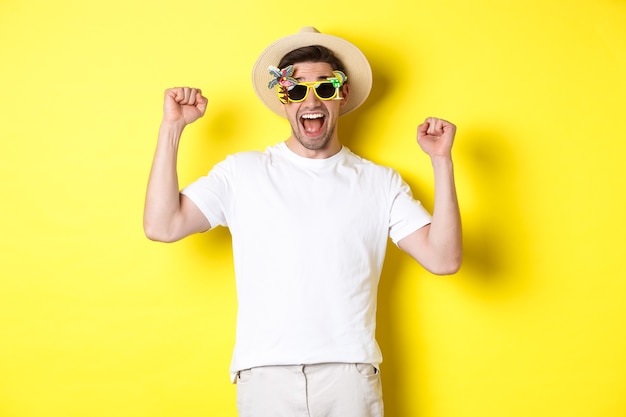 Pojęcie turystyki i stylu życia. Szczęśliwy człowiek wygrywający wycieczkę do kurortu, krzyczący tak i podnoszący ręce, triumfujący, noszący okulary przeciwsłoneczne i letni kapelusz, żółte tło.
