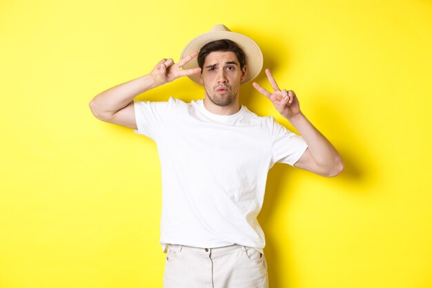 Pojęcie turystyki i wakacji. Fajny facet robiący zdjęcie na wakacjach, pozujący ze znakami pokoju i noszący słomkowy kapelusz, stojący na żółtym tle