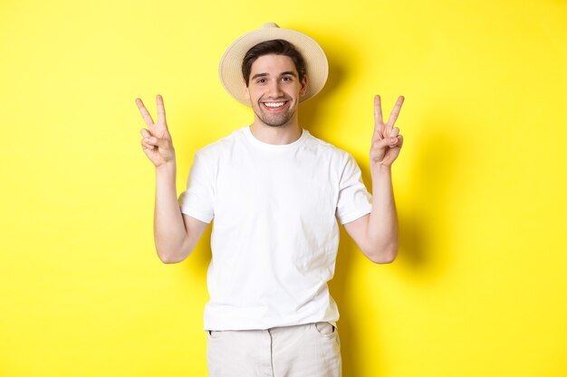 Pojęcie turystyki i wakacji. Szczęśliwy turysta mężczyzna pozuje do zdjęcia ze znakami pokoju, uśmiechając się podekscytowany, stojąc na żółtym tle.