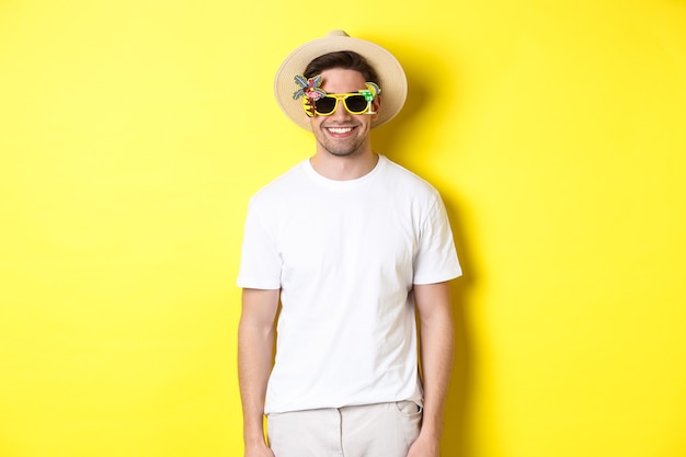 Pojęcie turystyki i wakacji. Zrelaksowany uśmiechnięty mężczyzna cieszący się kolacją w okularach przeciwsłonecznych i słomkowym kapeluszu, żółte tło.
