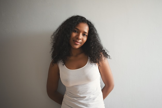 Bezpłatne zdjęcie pojedyncze ujęcie atrakcyjnej uroczej młodej afroamerykanki z obszernymi czarnymi włosami i czystą, idealną skórą, pozuje na pustej ścianie w białym podkoszulku, z nieśmiałym słodkim wyrazem
