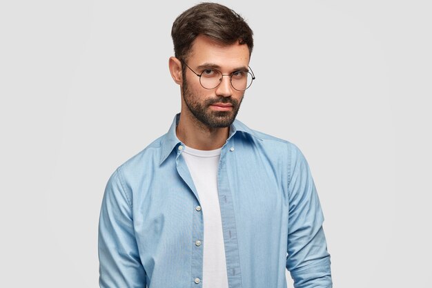 Pojedyncze ujęcie przekonany poważnych nieogolonych mężczyzn rasy kaukaskiej patrzy przez okrągłe okulary, ubrany w modną niebieską koszulę, odizolowane na białej ścianie. Ludzie, myśli, koncepcja stylu życia