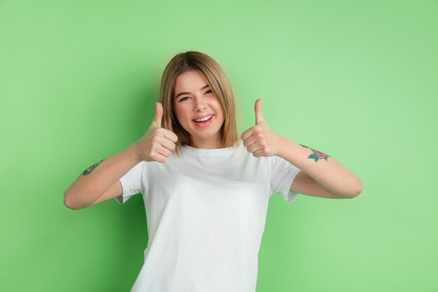 Pokazuje kciuki w górę. Kaukaski portret młodej kobiety na białym tle na ścianie zielone studio.