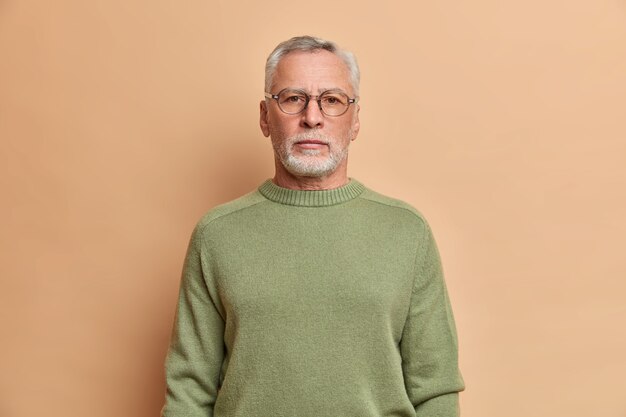 Półdługie ujęcie poważnego brodatego mężczyzny wygląda na pozbawionego emocji z przodu z surowym wyrazem twarzy, nosi okulary, a sweter ma siwe włosy, pewny siebie w czymś odizolowanym na beżowej ścianie studia