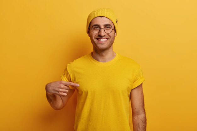 Półdługie ujęcie wesołego mężczyzny wskazuje na żółtą koszulkę, ma zadowoloną minę, reklamuje nowy strój, pozuje na jasnym tle