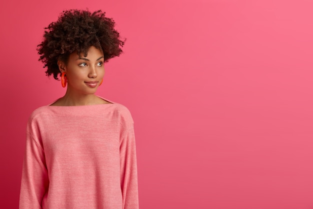 Półdługie ujęcie zamyślonej ciemnoskórej Afroamerykanki odwraca się, gdzieś skupiona, rozważa lepsze sposoby, ma coś na myśli, nosi sweter z długimi rękawami, odizolowany na różowej ścianie