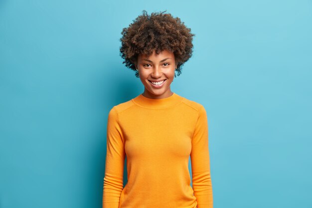 Połowa długości strzał całkiem wesoła młoda kobieta z ładnym promiennym uśmiechem zadowolona wyrazem ubrana w dorywczo pomarańczowy sweter na białym tle na niebieskiej ścianie