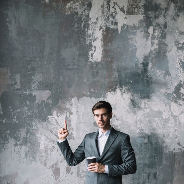 Pomyślna młoda biznesmen pozycja przeciw betonowej ścianie trzyma filiżankę kawy wskazuje jego palec oddolnego