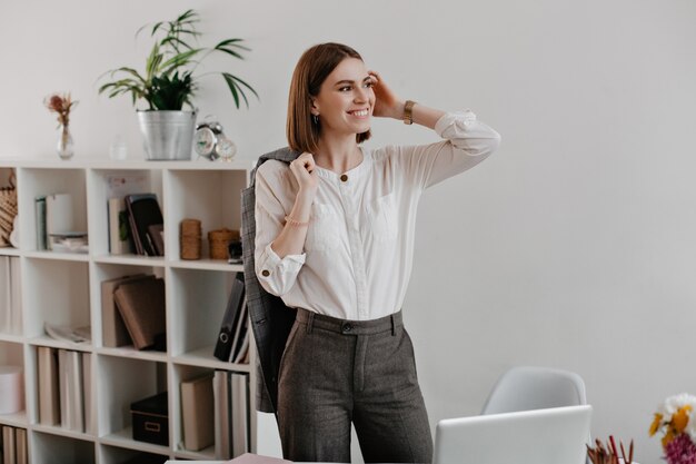 Pomyślna pracownica biurowa w szare spodnie i lekką koszulę z uśmiechem pozuje stojąc w miejscu pracy.