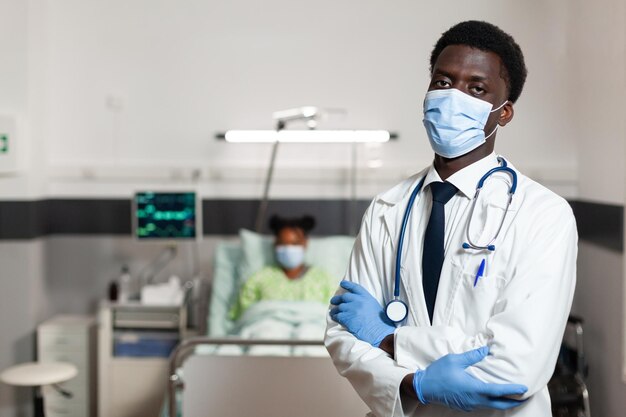 Portret afroamerykańskiego lekarza lekarza z maską ochronną, aby zapobiec zakażeniu covid19, patrząc w kamerę, stojąc na oddziale szpitalnym. Regeneracja pacjenta w tle