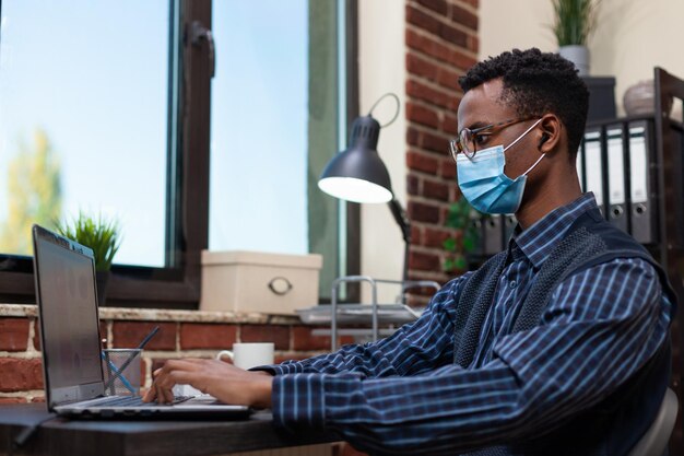 Portret afroamerykańskiego przedsiębiorcy, noszącego covid ochronną maskę na twarz, pracującego z analizą biznesową na laptopie. Pracownik biurowy zajmujący się uruchamianiem, analizujący wykresy sprzedaży, noszący ochronę przed wirusem koronowym.
