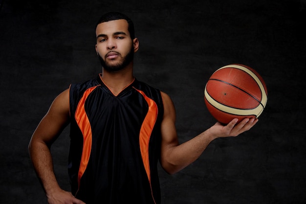 Portret afroamerykańskiego sportowca. Koszykarz w odzieży sportowej z piłką na ciemnym tle.