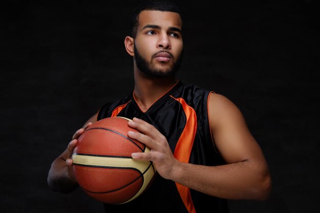 Portret afroamerykańskiego sportowca. Koszykarz w odzieży sportowej z piłką na ciemnym tle.