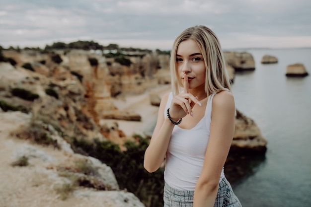 Portret atrakcyjna blondynka z gestem ciszy, pozowanie na kamienistej plaży z pięknem oceanu krajobraz.